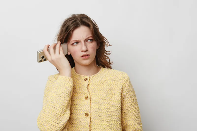Confused young woman in yellow sweater holds smartphone to ear, looking puzzled. Isolated on white background. Concept for misunderstanding, communication, and technology issues.