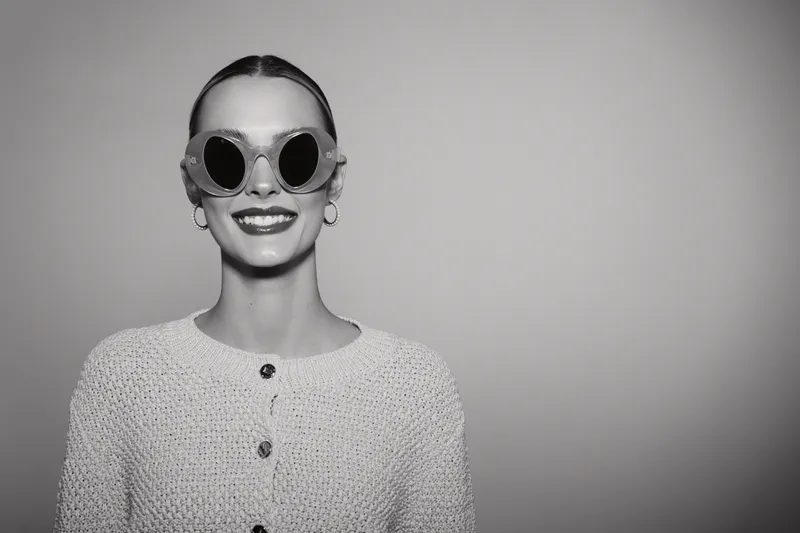 Smiling white woman with fair skin wearing round sunglasses and a light knitted sweater in a studio, portrait in black and white. Concept of fashion and style.
