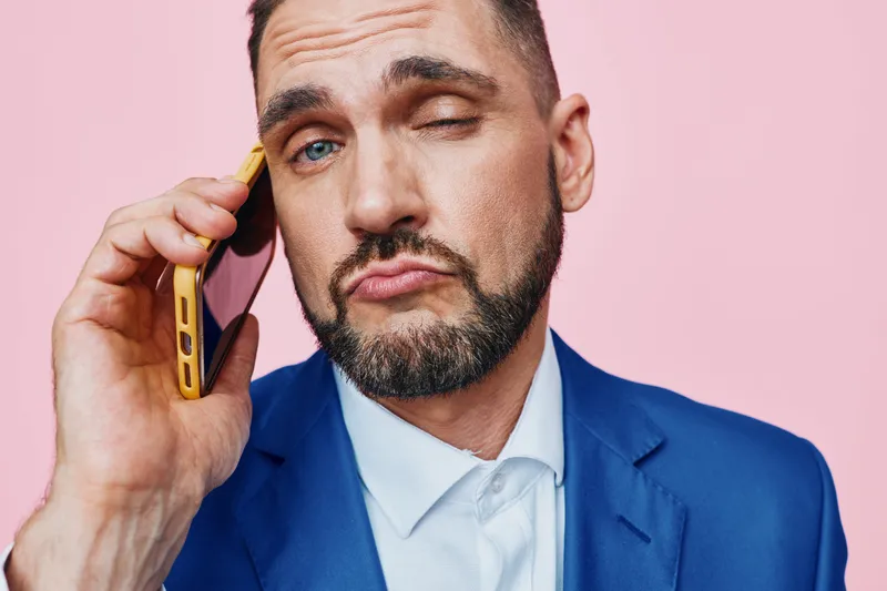 Portrait of a professional man in a blue suit on a solid pink backdrop, talking on a phone. The isolated colored background highlights a business mood, confident focus, and polished presentation.