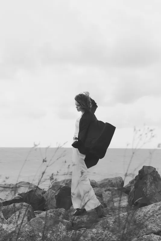 Woman walking along coastal rocks with flowing coat, windy sea breeze, monochrome scenery, dramatic seascape conveying solitude, travel mood, outdoor fashion