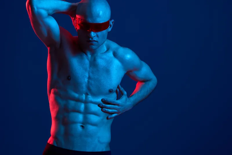 Confident man posing with futuristic sunglasses, showcasing a strong physique. The striking blue lighting emphasizes his muscular features in this vibrant color image.