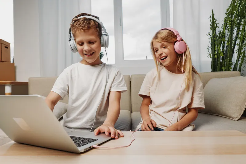 Two happy children wearing headphones enjoy music and laughter together while sitting on a couch in a bright living room with a laptop on the table. Concept of friendship and joy.