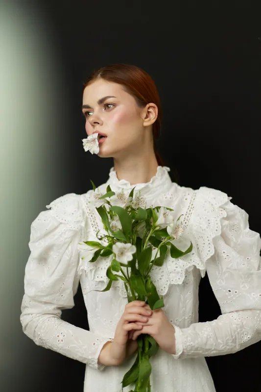 Elegant young woman with a bouquet of flowers, wearing a vintage white dress, posing against a dark background, evoking feelings of grace and beauty in a seasonal setting