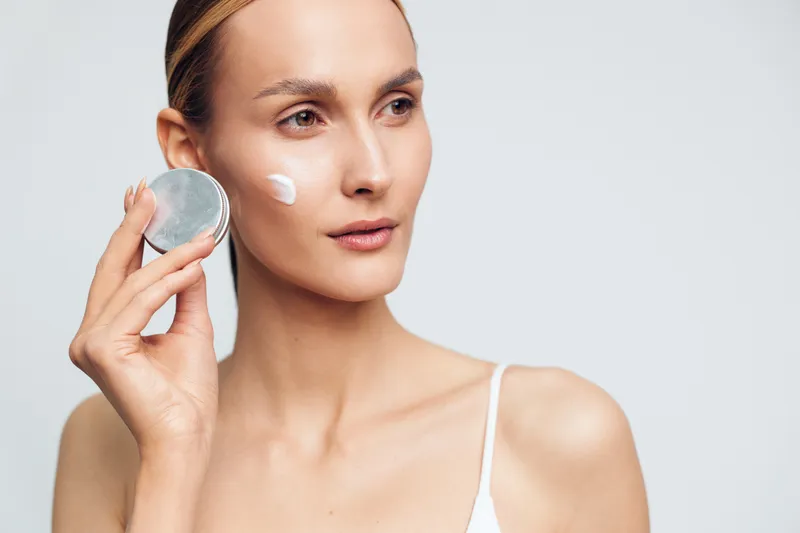 Young woman with clear skin applying white face cream on cheek using a small jar. Close-up portrait on a light background. Concept of skincare, beauty, and self-care routine.