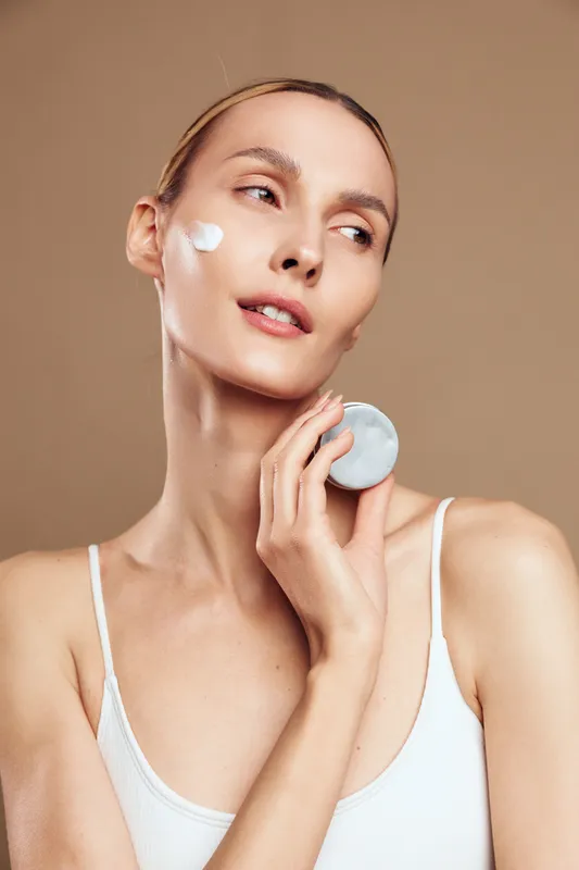 A young woman in a white tank top applies cream to her cheek using a cotton pad against a neutral background. Concept of skincare, beauty routine, and self-care.