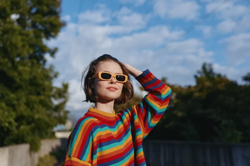 Woman in a rainbow sweater and yellow sunglasses, young adult with short hair smiling outdoors, leaning back with hand in hair in suburban sunlight, casual outfit, confident vibe.