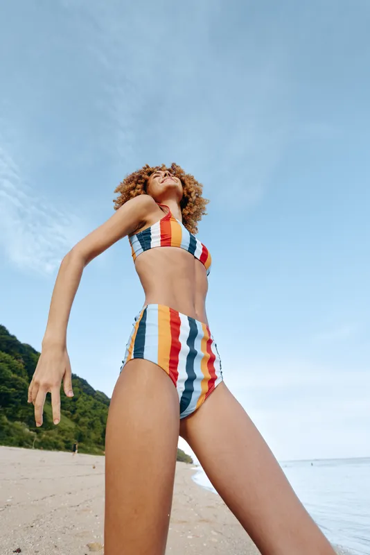 Beach woman bikini summer ocean sky outdoors vacation, striped swimwear on sunny shoreline, woman with curly hair posing in profile, showing legs and body, soft daylight mood, travel relaxation