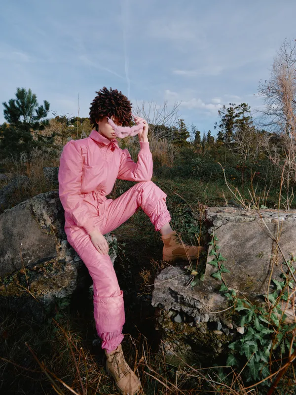 Pink jumpsuit clad model posing on a rocky ledge outdoors, sunlit scenery, thoughtful gaze, rugged landscape, fashion moment captured in natural light with earthy tones and calm mood.