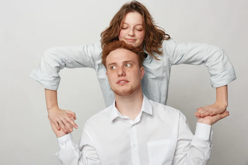 Young man and woman in white shirts pose together on light background. The woman stands behind the man holding his hands. Concept of friendship, support, and casual style.