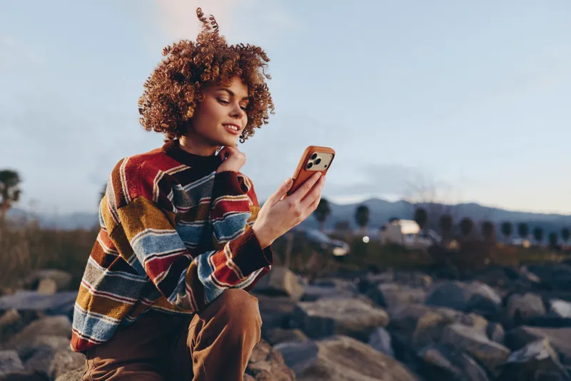 Woman lifestyle outdoors, curly hair and plaid sweater, sitting by rocky shore, smiling while using smartphone, sunset light, relaxed casual mood, calm waves and sky