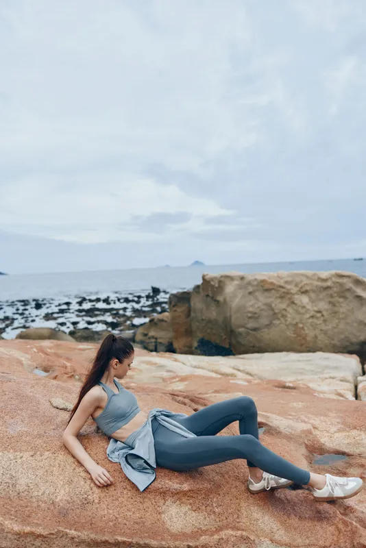 Woman practicing yoga on a rocky beach at sunset, wearing stylish workout clothes, conveying peace and focus in a serene coastal setting