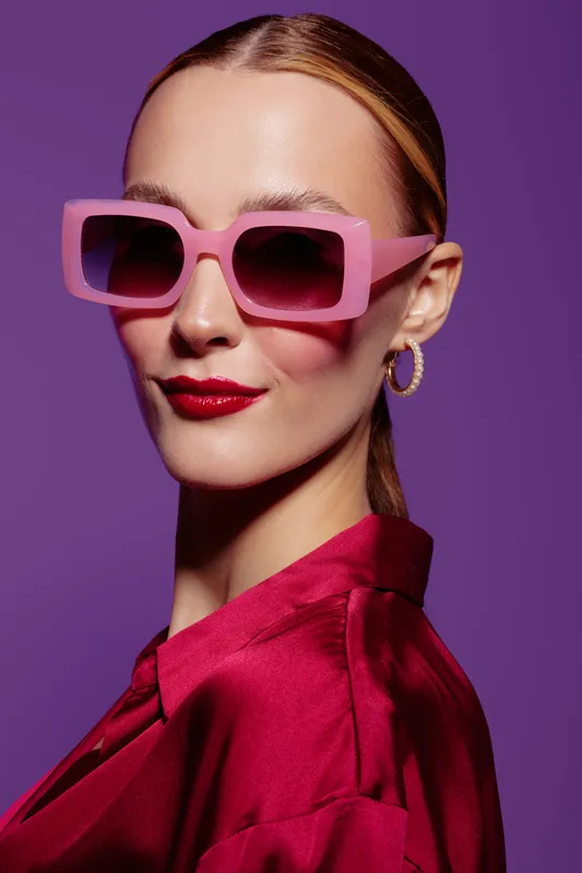 Stylish woman with pink sunglasses and red lipstick poses confidently against purple background. Fashion portrait with vibrant colors and modern accessories for beauty and style concept.