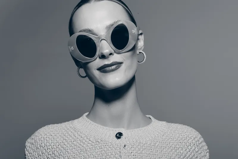 Stylish caucasian woman with fair skin and slicked hair wearing round sunglasses and knitted sweater poses confidently in studio. Concept of fashion, elegance, and modern style.