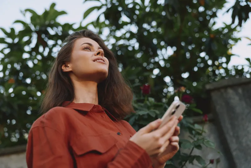 A woman in a rust red shirt stands outdoors in a garden, looking up with a thoughtful gaze while holding a smartphone. Warm natural light conveys casual, reflective lifestyle
