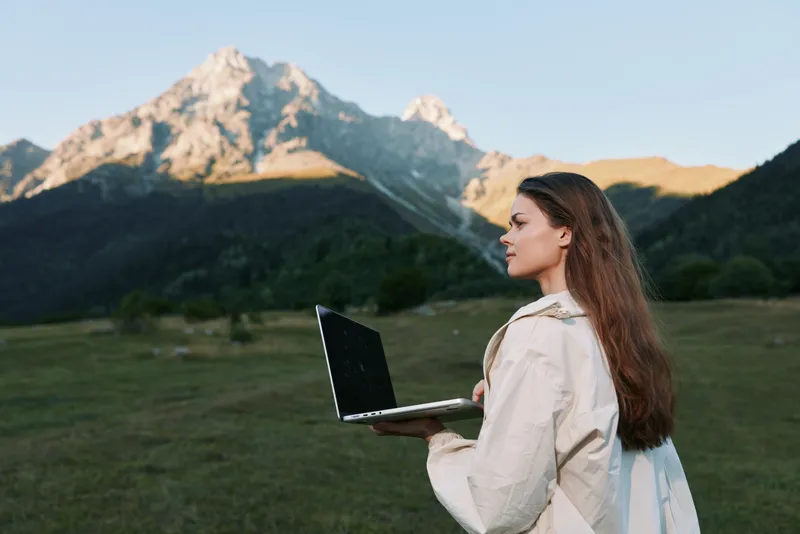 A focused young woman stands outdoors with a laptop in a remote landscape, ready to work. She appears calm and confident, blending technology with nature to convey productive, adaptive work in open