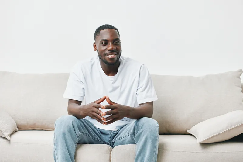 Smiling African American man sitting on a sofa in casual white t shirt and jeans, exuding positivity and warmth in a bright, minimalist living room