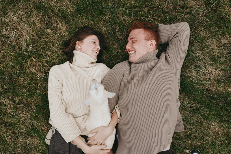 A joyful couple lies on the grass in cozy sweaters, smiling at each other while a white goat kid rests between them. An intimate lifestyle moment outdoors with animals, warmth, and simple happiness.