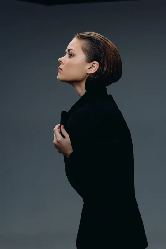 Profile of a serious woman in dark clothing with slicked-back hair against a gray background