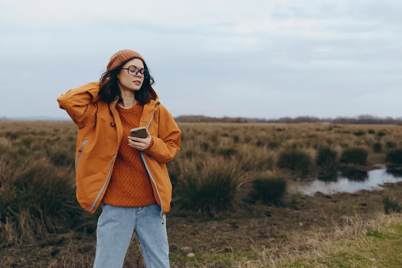 Woman lifestyle outdoors in nature by a river in autumn, orange jacket and glasses, relaxed walk, calm mood, wide landscape background, soft daylight