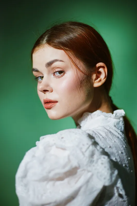 A young woman with shoulder length brown hair in a vintage white blouse poses against a soft green background, exuding a thoughtful and serene expression