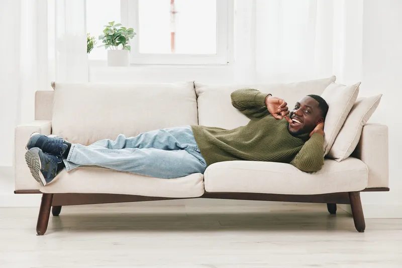 Happy black man relaxing on a comfortable sofa, enjoying leisure time and smiling, dressed in a cozy sweater and jeans, bright interior