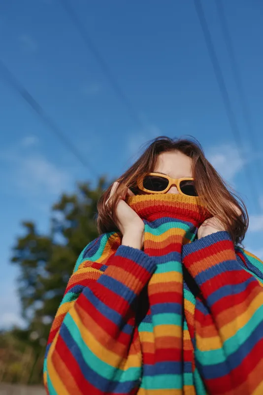 Woman in a colorful striped sweater and yellow sunglasses outdoors, young adult posing playful portrait in a casual outfit, covering face with cozy knit pullover and joyful eyes.