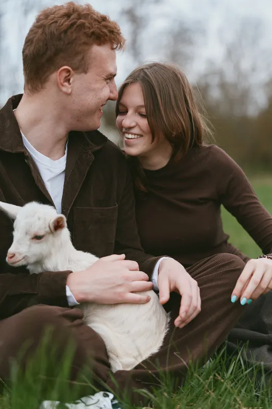 Candid couple smiles while gently holding a playful goat kid in an open field, capturing rustic lifestyle, bonding, and care. Natural daylight, soft tones, warm mood, outdoor companionship.