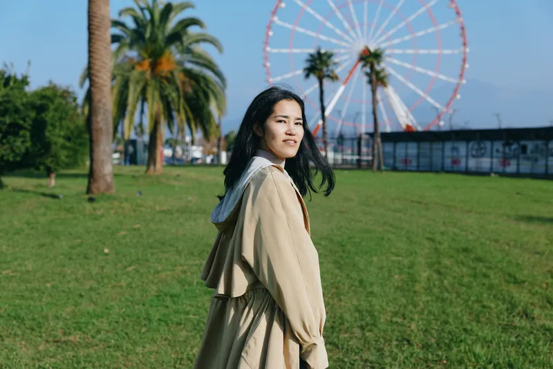 Young woman in beige coat strolling through a sunny park with a large ferris wheel in the background, casual fashion, cheerful mood, daylight, open grass, palm trees, gentle breeze.