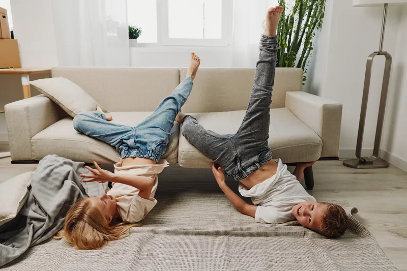 Two children playfully lying on the floor with legs raised in a bright living room. Casual home environment with natural light, carpet, sofa, and relaxed joyful mood.