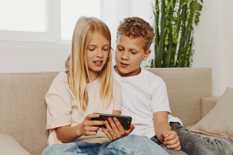 Two children sitting on couch watching smartphone together, sharing content and enjoying modern digital entertainment indoors.