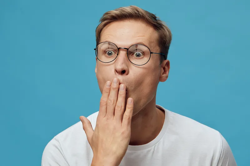 surprised caucasian man wearing glasses white t-shirt covering mouth with hand isolated on blue background facial expression reaction concept digital detox
