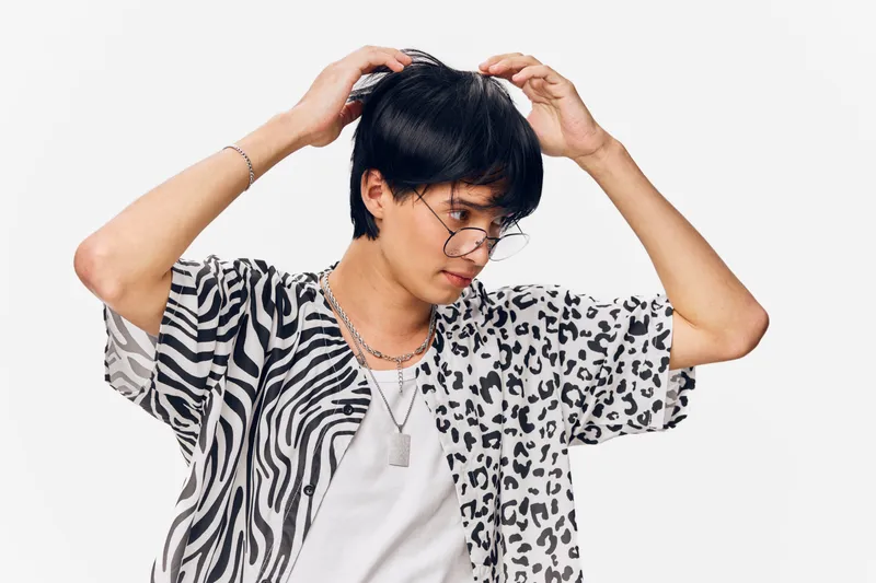 young man wearing mixed animal print shirt and round glasses adjusting hair against white background