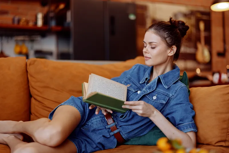 Женщина отдыхает на диване, читает книгу, на ней джинсовая рубашка и джинсы