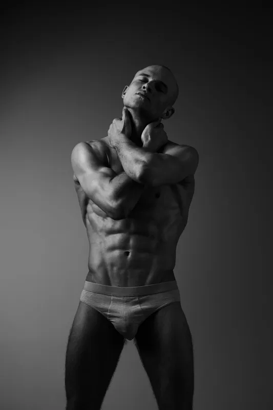 Black and white portrait of a muscular man posing with confidence, showcasing strong physique and dramatic lighting that highlights contours and shadows.