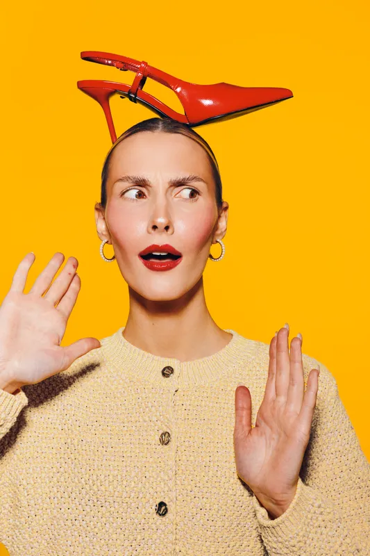Surprised woman with red high heel shoe balanced on her head, wearing beige sweater and gold hoop earrings. Bright orange background creates vibrant and playful mood.