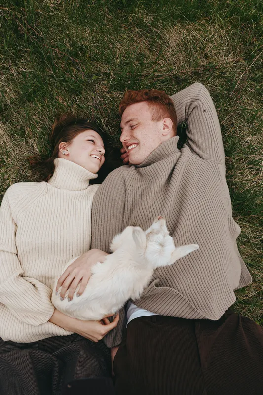 Joyful couple lying on grass with a playful goat kid, cozy sweaters, intimate moment, relaxed lifestyle scene, natural light, outdoor romance, candid harmony.