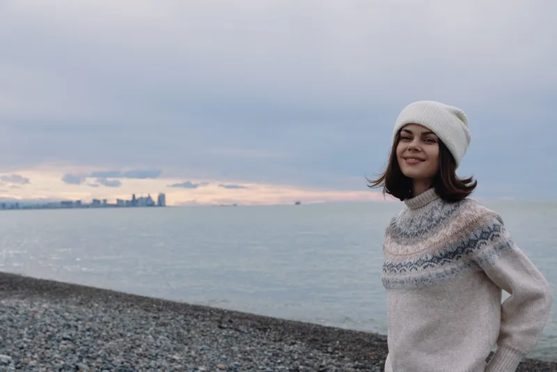 Woman in a knitted sweater and beanie stands on a pebbled beach by the calm sea at sunset, smiling with a distant skyline on the horizon.