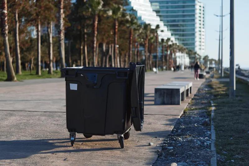 A large black waste bin on a sunny urban promenade stands beside the waterfront with distant pedestrians and tall modern buildings in the background, creating a candid street scene of everyday city