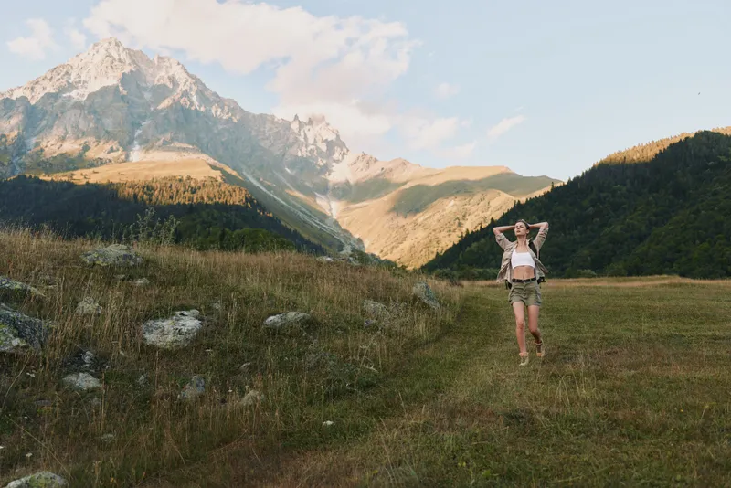 Young woman stands in a grassy meadow with towering mountains in the background, looking relaxed and confident.