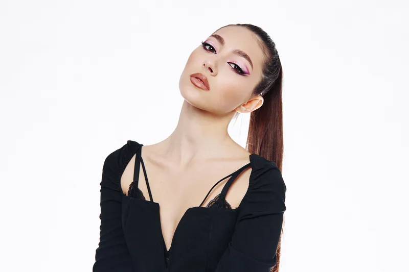 Fashion model with sleek ponytail and dramatic purple eyeliner poses confidently in black trendy outfit. White studio background highlights beauty and edgy style concepts.