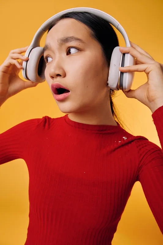 Young woman in red sweater wearing headphones and listening to music with surprised expression on yellow background. Female person enjoying audio sound with casual style and studio lighting.