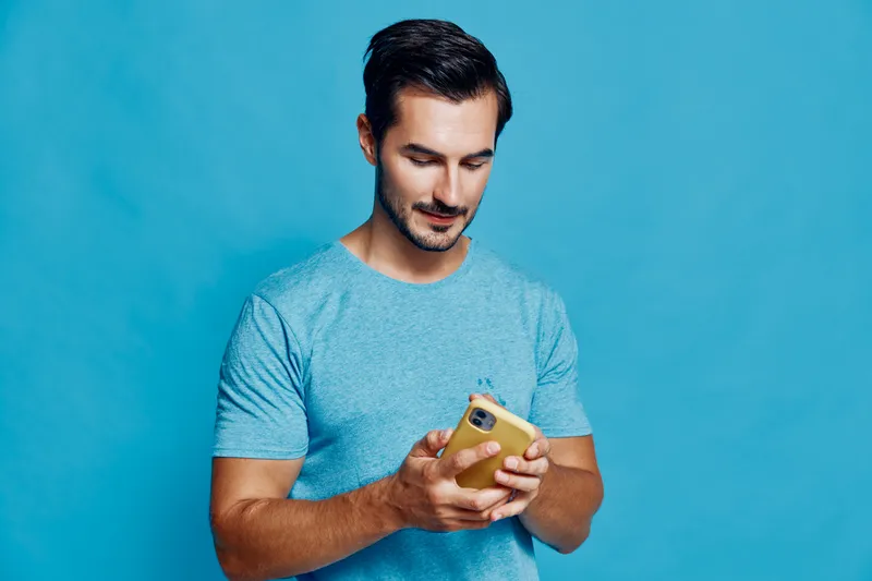 Young man wearing a blue t-shirt smiles while using a mobile phone, focused and engaged in conversation isolated against a vibrant solid blue background.