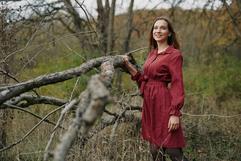 A candid outdoor portrait of a woman in a red dress standing near a fallen tree in a quiet forest, capturing authenticity and genuine mood under soft, natural light.