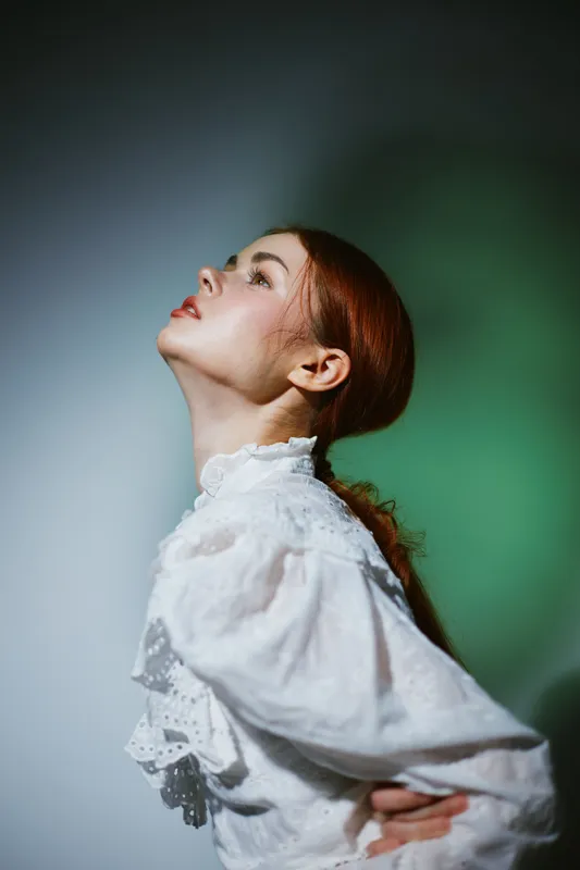 Portrait of a young woman with red hair, wearing a white vintage blouse, expressing contemplation against a softly lit green background