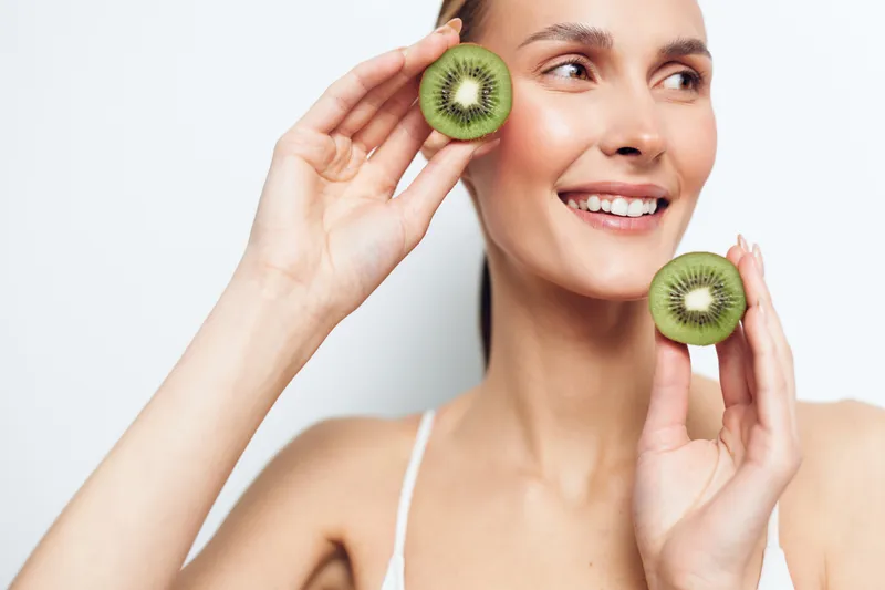 Woman skincare kiwi fruit beauty wellness fresh portrait concept, smiling young female holds kiwi slices on cheeks, bright studio lighting, natural face mask, clean white background