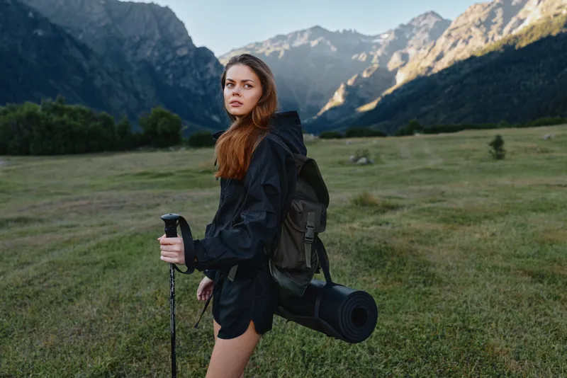 A confident hiker stands in a grassy valley with rugged mountains behind, carrying a backpack and a rolled mat. She looks forward with calm focus, ready for a rewarding outdoor journey.