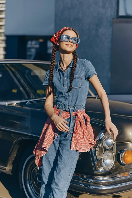 Lifestyle woman in modern boho-western denim street style with braids and silver sunglasses, smiling confidently in bright film color outdoors near classic car.