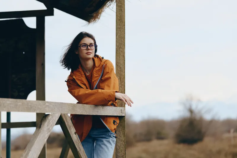 Woman lifestyle outdoors, wearing orange jacket and glasses, leans on the wood ladder, arms crossed, portrait pose. Rural field and distant trees in soft daylight create a relaxed country fashion