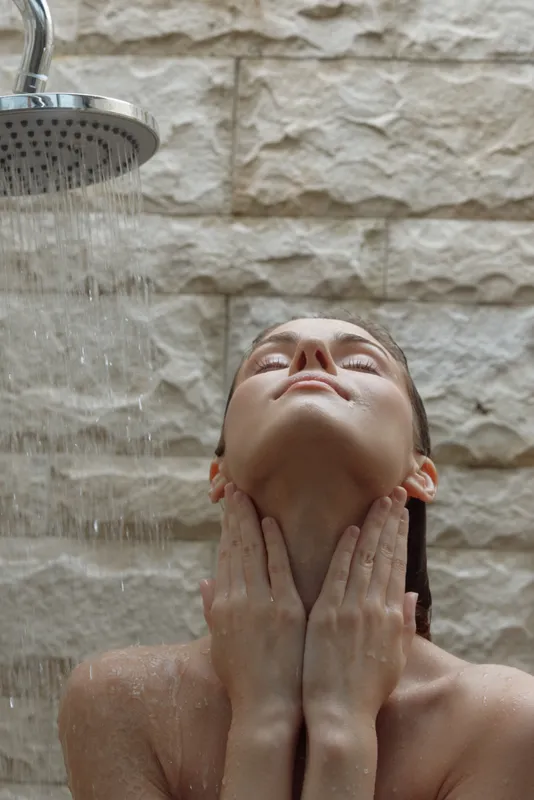 Woman enjoying relaxing shower with water falling on her face against textured stone wall, representing tranquility and self-care in a natural bathroom setting.