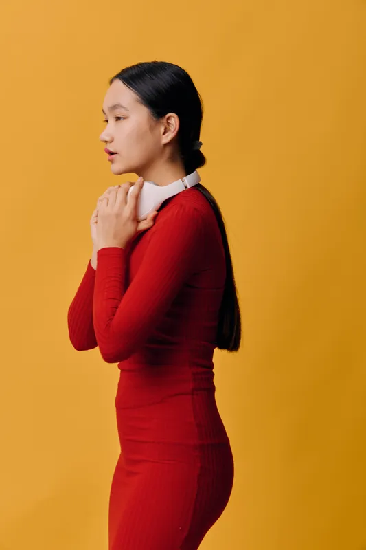 Young woman in red outfit standing sideways holding headphones on yellow background. Casual style with long hair tied back and minimal makeup for lifestyle concept.